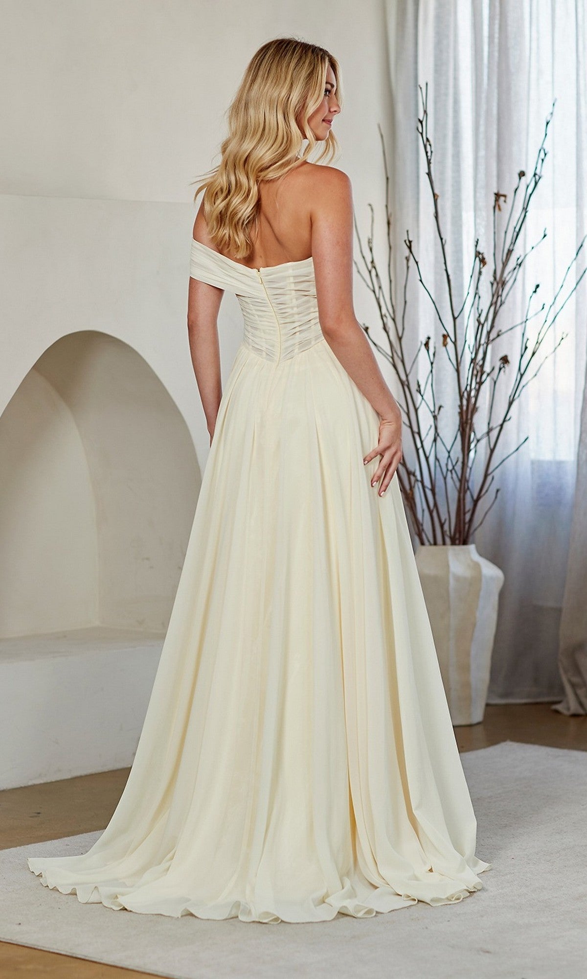 A woman wearing the Amelia Couture 9097 long prom dress stands indoors with her back to the camera, near a vase of bare branches and sheer curtains.
